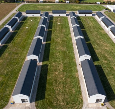 An aerial view of a clean, structured, and modern breeding facility. The layout is perfectly symmetrical, showing green paddocks and sleek dark-colored shelters, representing the precision of the DORPRIME integrated system.