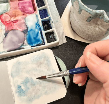 A person holding a paintbrush painting in a skechbook with a watercolor palette in the background.