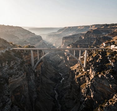 A breathtaking drone shot looking down at a bridge spanning a deep canyon in a Global / International landscape. The composition is symmetrical and cinematic, with soft morning light and a slight mist, incorporating a soft off-white and deep charcoal color profile.