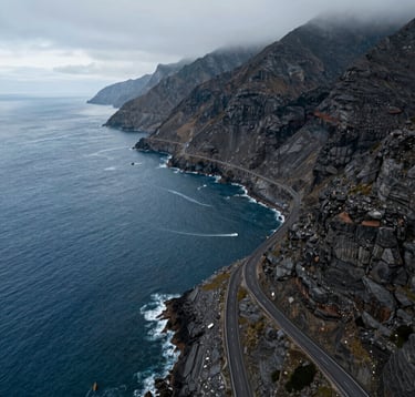 An aerial drone photograph of a winding coastal road in a Global / International location, with the deep blue ocean on one side and jagged cliffs on the other. Cinematic lighting with a soft grey atmospheric mist and charcoal rock textures.