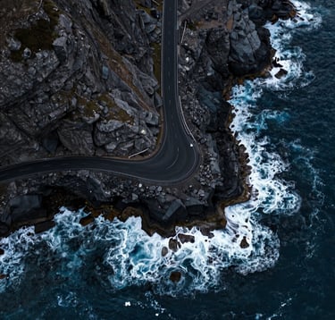 A high-altitude drone shot looking straight down at a rugged coastline with a single road hugging the cliffs. The deep blue water contrasts with the Midnight Black asphalt. Soft Pearl foam from the waves creates a dynamic pattern. Cinematic aerial photography in a Global / International location.
