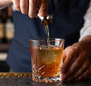 Extreme close-up of a mixologist’s hands using a glass smoker on a vibrant amber cocktail in a heavy crystal glass. Dark slate blue and charcoal background with gold light. Urban luxury bar setting.