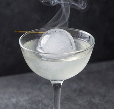 Close-up photography of a transparent cocktail glass featuring a large, clear ice sphere and a minimalist garnish. Wisps of smoke from dry ice drift across a dark charcoal background. North American / Urban.