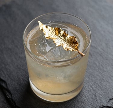 Macro photography of an exotic cocktail being garnished with a branded edible gold leaf, deep shadows and slate grey textures, cinematic focus in a North American / Urban lounge.