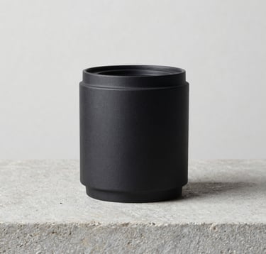 A refined still life photograph of a single charcoal black design object resting on a light gray stone surface. The background is a clean off-white wall in a Northern European studio. The lighting is precise, casting a soft medium gray shadow, embodying a modern and professional aesthetic.