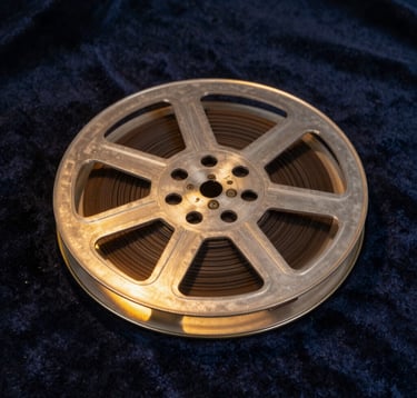 A close-up of a vintage film reel canister sitting on a dark navy velvet surface, illuminated by a sharp gold spotlight from the side. The metallic edges catch the light, suggesting a fusion of history and modern luxury.