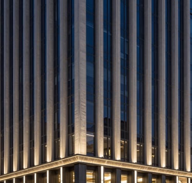 The sleek architectural facade of a luxury contemporary building in Seoul at night. The structure features clean vertical lines and is illuminated by a subtle deep navy and soft gold lighting scheme. The composition is clean and modern, evoking high-end urban elegance.