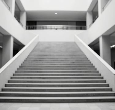 A black and white architectural detail of symmetrical stairs leading to a high-ceilinged atrium. The composition is clean and analytical, highlighting structure and order. Hints of brand color #94A7B7 in the shadows.