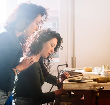 Dos artesanas trabajando juntas en un banco de taller de joyería iluminado por el sol 