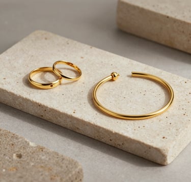 An elegant, understated luxury still-life photograph featuring delicate gold jewelry, including rings and a bracelet, resting on natural stone slabs of soft beige and taupe colors. Soft directional lighting highlights the polished gold textures. Calm, minimal, editorial design style. Global / North American and Western European.