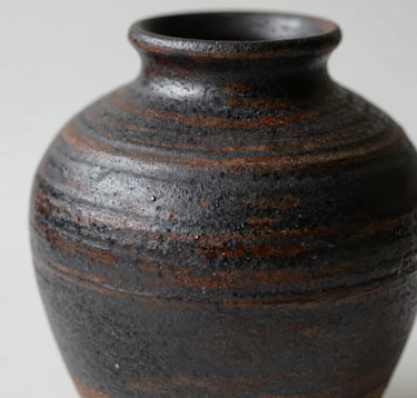 Close-up of ceramic textures featuring dark charcoal (#1A1A1A) and earthy brown (#5B4B3A) glazes. The lighting is dramatic, showcasing the tactile, rustic surface of a handmade vase against a minimalist background.