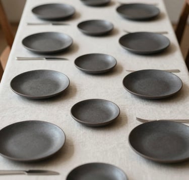 A top-down composition of a minimalist table setting for a tasting menu. A series of small ceramic plates in dark tones (#1A1A1A, #5B4B3A) arranged symmetrically on a light linen cloth, reflecting an elegant gallery aesthetic.