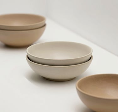 A minimalist gallery-style photograph of the 'Serie Alba' collection. Three stacked ceramic bowls with organic edges in varying shades of sand (#EAE4D3) and tan (#C2BEB0). Clean shadows, white museum-like environment.