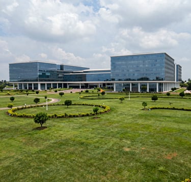 A wide shot of an expansive, green technology park in Bangalore. The buildings are futuristic with glass facades and Pearl White steel columns. The setting reflects a high-tech South Asian / Indian innovation hub with sustainable landscaping.