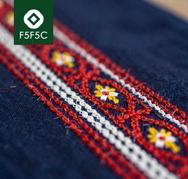 A close-up, high-detail macro shot of traditional Dao ethnic embroidery. The intricate patterns of red, white, and yellow thread on dark indigo fabric tell a story of heritage. Soft focus background featuring the cooperative's logo. Colors emphasize #1A3D2F and #F5F5EC.
