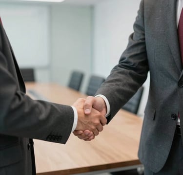 Close-up photography of two professionals in dark slate grey suits shaking hands in a bright, modern boardroom. A hint of crimson red is visible in a tie. The atmosphere is professional and trustworthy.