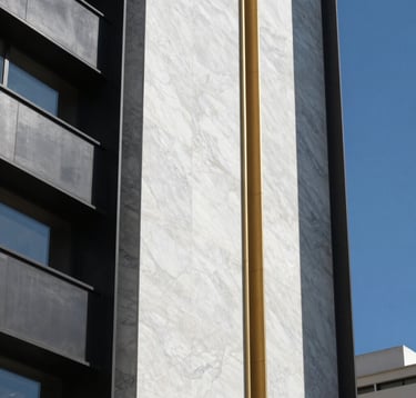 A detail shot of high-end architectural materials: a combination of white marble, black steel, and gold finishings on a building facade. Set against a clear blue sky in a high-altitude South American / Ecuadorian city, conveying minimalist luxury.