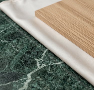 A close-up, artistic shot of interior design textures. Features a slab of royal dark green marble with intricate white veining, layered with soft off-white silk fabric and a sample of warm sandy beige oak wood.