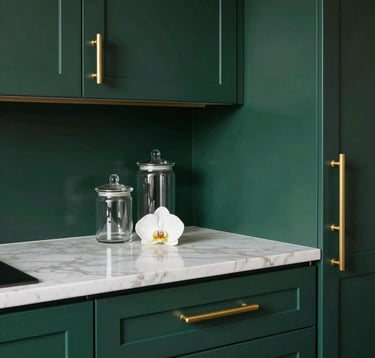 A close-up architectural shot of a luxury modular kitchen. The cabinets are finished in a deep royal dark green with brushed metallic gold handles. The countertop is a thick slab of white marble with subtle beige veining. Elegant glass jars and a single orchid sit on the counter.