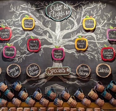 Hand-drawn chalkboard menu featuring hard cider flight flavors and specials above a row of ceramic mugs.