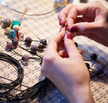 Manos artesanas con uñas rojas elaborando un collar hecho a mano con cuentas de vidrio moradas