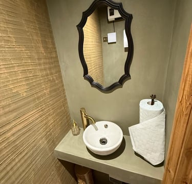 Rustic bathroom with white washbasin, gold tap and black mirror set against olive green microcement