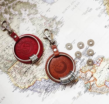 Red and brown leather Vagabond Life keychains with engraved travel rings on a vintage world map.