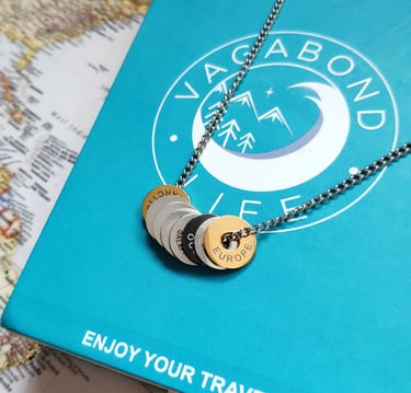 Vagabond Life engraved continent rings on a silver chain necklace over a blue travel box and world map.