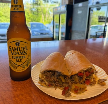 A Samuel Adams Summer Ale beer bottle next to a loaded Philly cheesesteak sub with peppers on a paper plate.