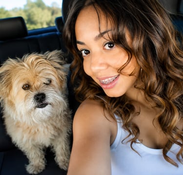 A smiling young woman with curly hair takes a car selfie with her small, fluffy tan dog.