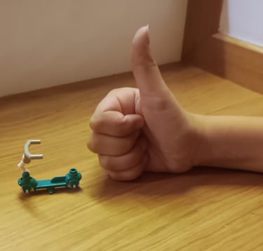 Dłoń dziecka z podniesionym do góry kciukiem, na biurku, obok maleńkiej hulajnogi z klocków.