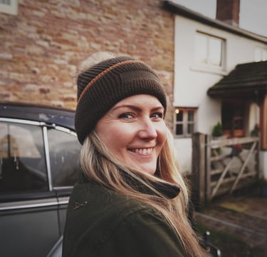 Smiling woman in a winter bobble hat and green coat standing by a vintage car in front of a rustic stone house.