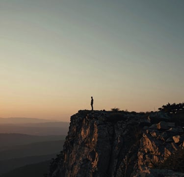 A wide-angle, cinematic photograph of a serene landscape at twilight. A lone figure stands in the distance on a rugged cliff, bathed in the soft, warm glow of a setting sun. The lighting is nostalgic and soft, with deep shadows (#262626) and warm highlights (#F5F0E1, #C4B29A). The composition evokes a sense of timeless elegance and emotional depth.