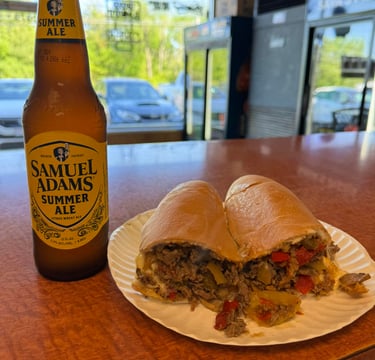 A Samuel Adams Summer Ale beer bottle next to a loaded Philly cheesesteak sub with peppers on a paper plate.
