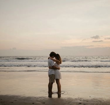 A warm, intimate wide shot of a couple embracing on a quiet Bali beach at dusk. The lighting is soft and golden, with the sand reflecting tones of #C8B6A6 and #F8F5EF. The composition is minimalist, emphasizing the emotional connection and the natural beauty of the island.