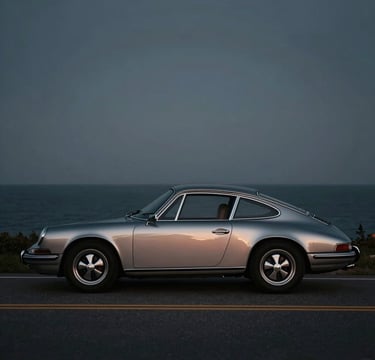 A side-profile shot of a vintage 1960s Porsche 911 in a metallic silver finish, parked on a coastal road at twilight. The lighting is cinematic, with soft highlights reflecting #B2BEB5 tones off the curves. The deep shadows of the background blend into the #1C2833 palette, emphasizing a sophisticated and timeless atmosphere.