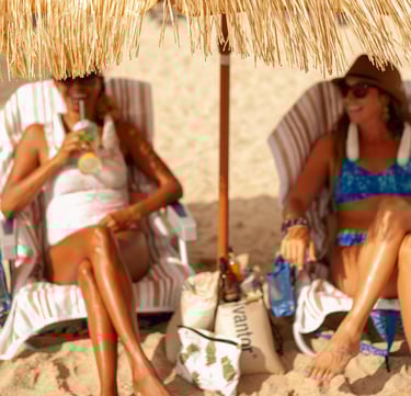 two women sitting on a beach under a straw umbrella