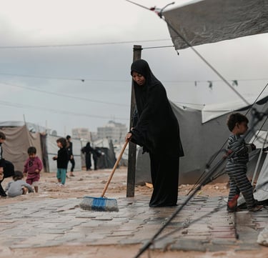 femme réfugiés gaza devant sa tente en train de nettoyer le sol, ses enfants jouent à l'extérieur dans la boue
