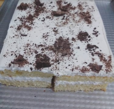 Layered vanilla sponge cake with white frosting and dark chocolate shavings on a baking sheet.