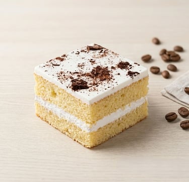 Square slice of vanilla sponge cake with white frosting and cocoa powder topping near coffee beans.
