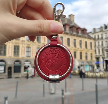 Red leather Vagabond Life travel keychain with engraved rings held in a city square.
