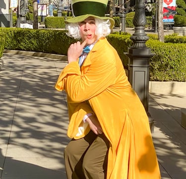 A Mad Hatter inspired performer with a large green top hat and yellow coat poses outdoors.
