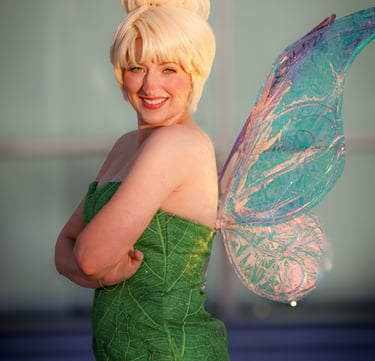 A fairy perfromer smiling featuring a green leaf dress and shimmering iridescent fairy wings.