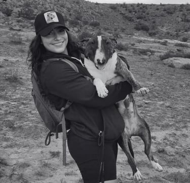 Mujer sonriente con mochila caminando al aire libre mientras sostiene a su Bull Terrier atigrado.