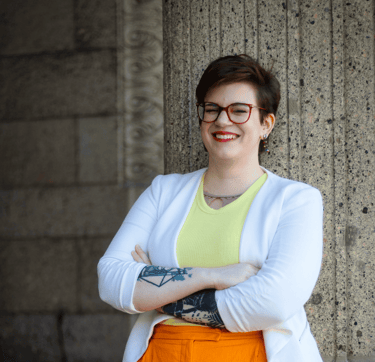 ein Foto von Swana Schuchmann. Sie trägt einen weißen Blazer und lehnt sich an eine Säule, lachend.
