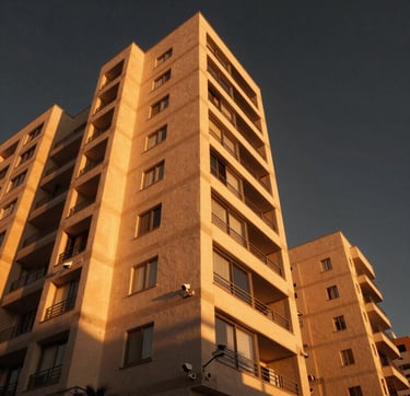 A cinematic, low-angle wide shot of a modern luxury residential building in a South American / Brazilian neighborhood during the golden hour. The building is illuminated by warm, sunny lighting, and subtle, professional electronic security cameras are visible on the architecture. The atmosphere is cozy yet secure, featuring vibrant orange tones from the sunset against a charcoal black sky.
