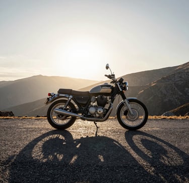 A wide-angle, cinematic photograph of a classic motorcycle parked on a high mountain pass in a Global / Western landscape. The lighting is low-sun golden hour, casting long shadows of deep charcoal across the pavement. The sky is a gradient of frost white and silver grey. Sophisticated and adventurous mood.