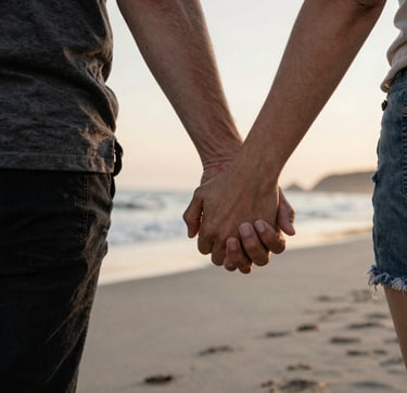 A candid, authentic photograph of a couple's hands interlaced while walking on a North American / US beach at sunset. The lighting is soft and sun-drenched. Hints of charcoal and warm brown appear in their clothing, contrasting softly with the sand and coastal water.