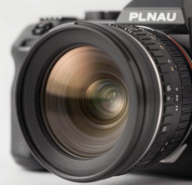 A close-up macro photograph of a professional cinema lens reflecting a soft, warm light from a high-end North American / US studio environment. The palette features muted champagne gold reflections against a dark slate grey and deep charcoal black camera body. The lighting is sophisticated and highlights the technical mastery and premium quality of the gear.