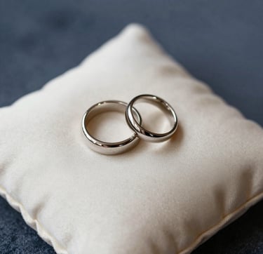 A macro photograph focusing on the fine details of a luxury wedding: a pair of bespoke rings resting on a soft cream silk cushion. The lighting is soft and ethereal. Background includes hints of deep navy blue and slate gray-blue textures. Sophisticated and minimalist composition, Hispanic setting context.